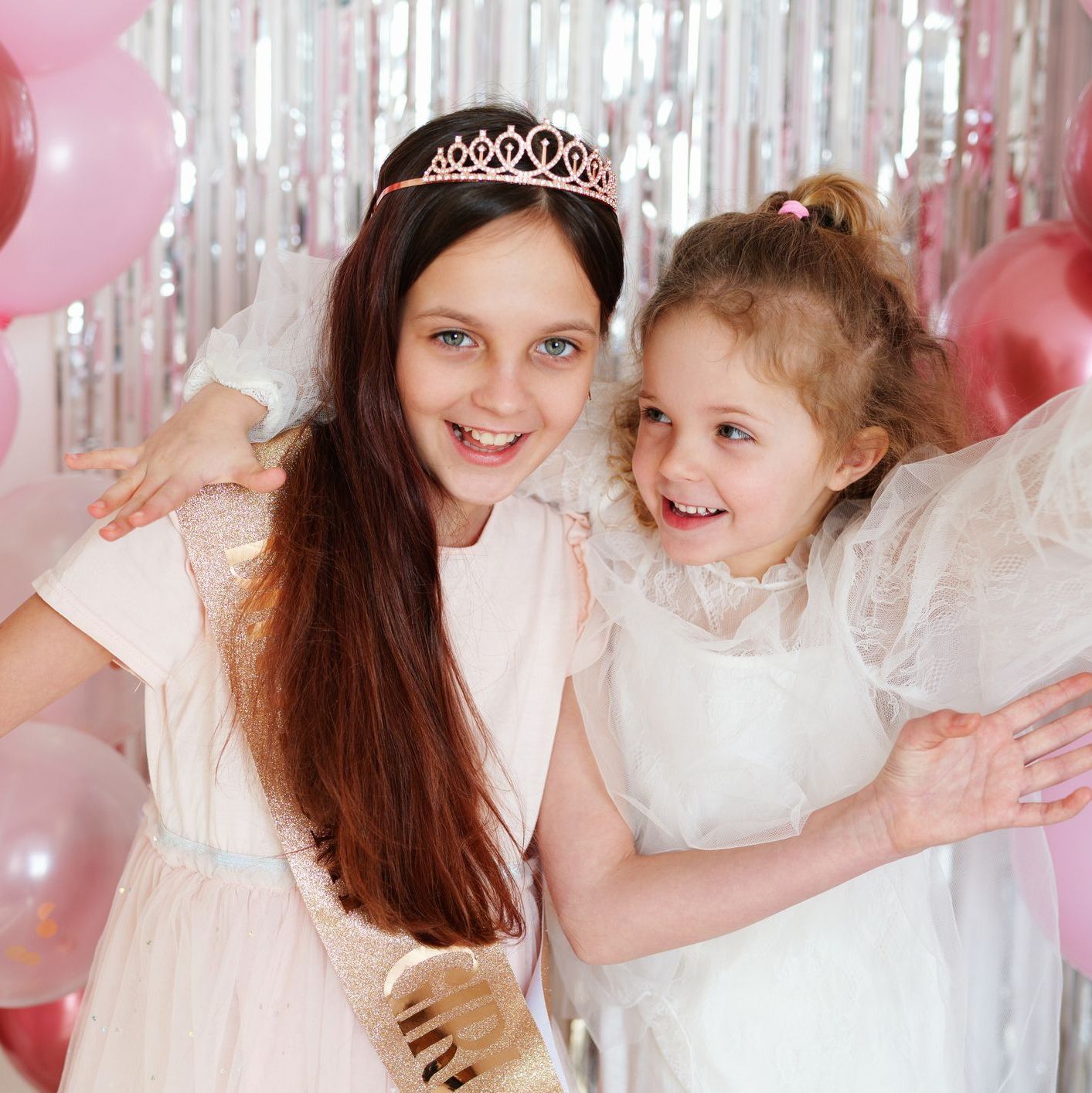 Two girls are having fun at a birthday party with pink balloons and decorations around them. They are smiling and posing happily, enjoying the celebration.